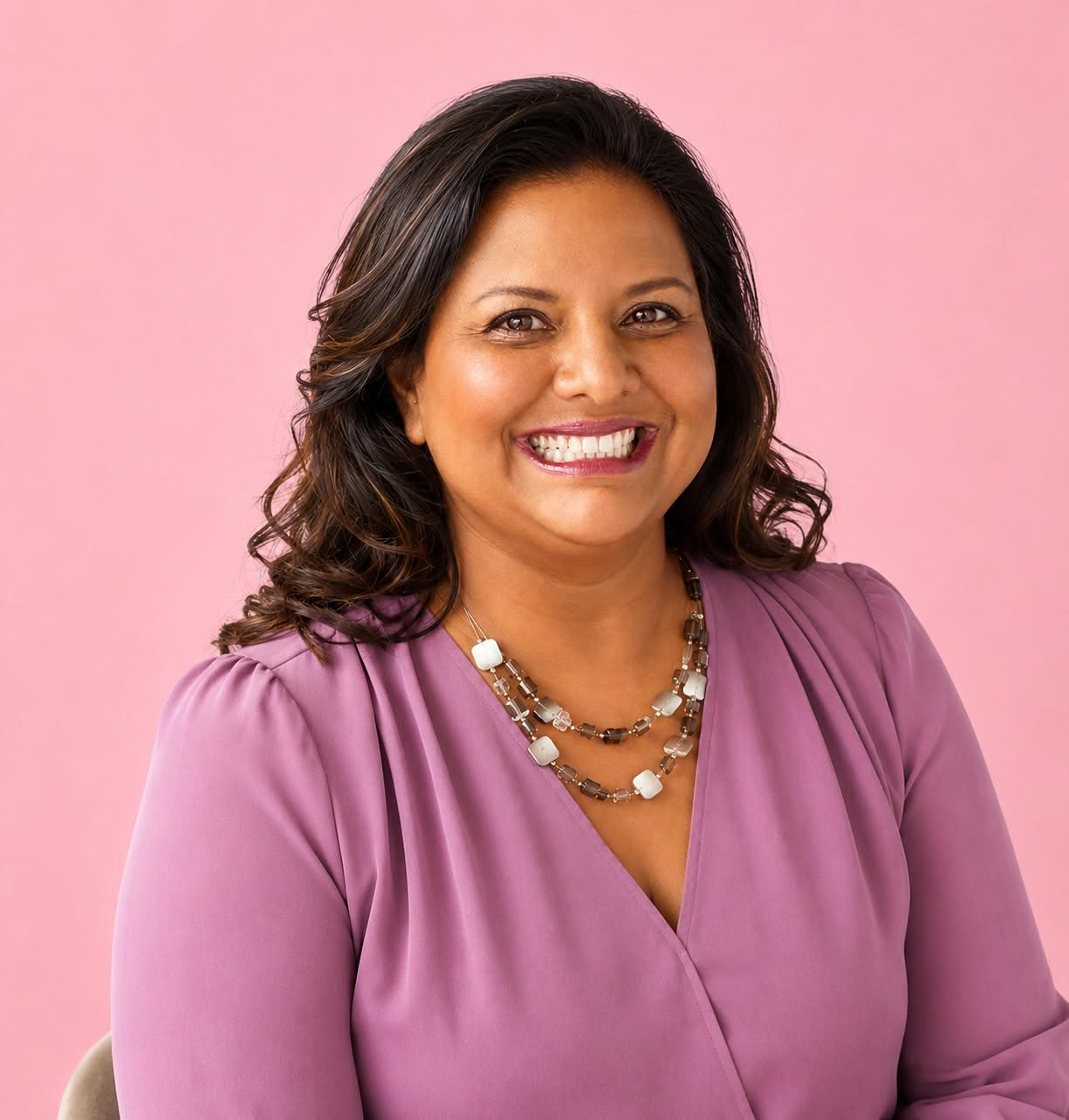 Smiling BusiWomen founder Karen Baring in a purple blouse and layered necklace against a pink background